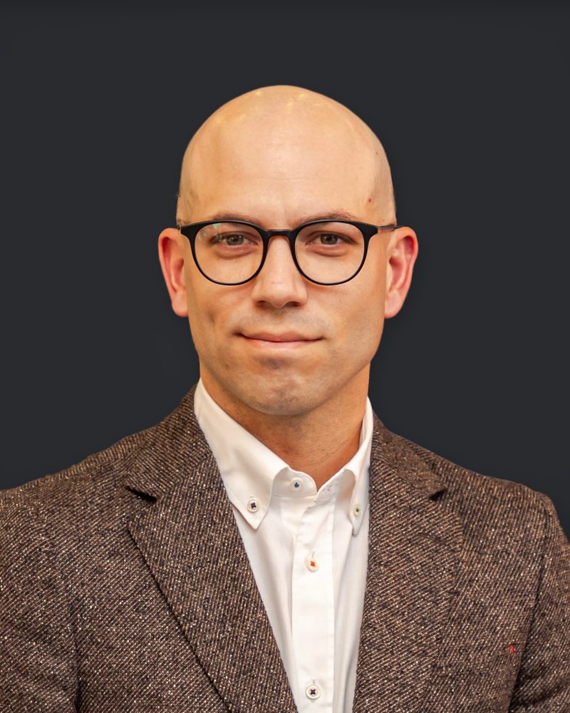Josué González, head-and-shoulders portrait. He smiles slightly and wears round black glasses, a white button-down shirt and a brown tweed jacket, against a plain charcoal background.